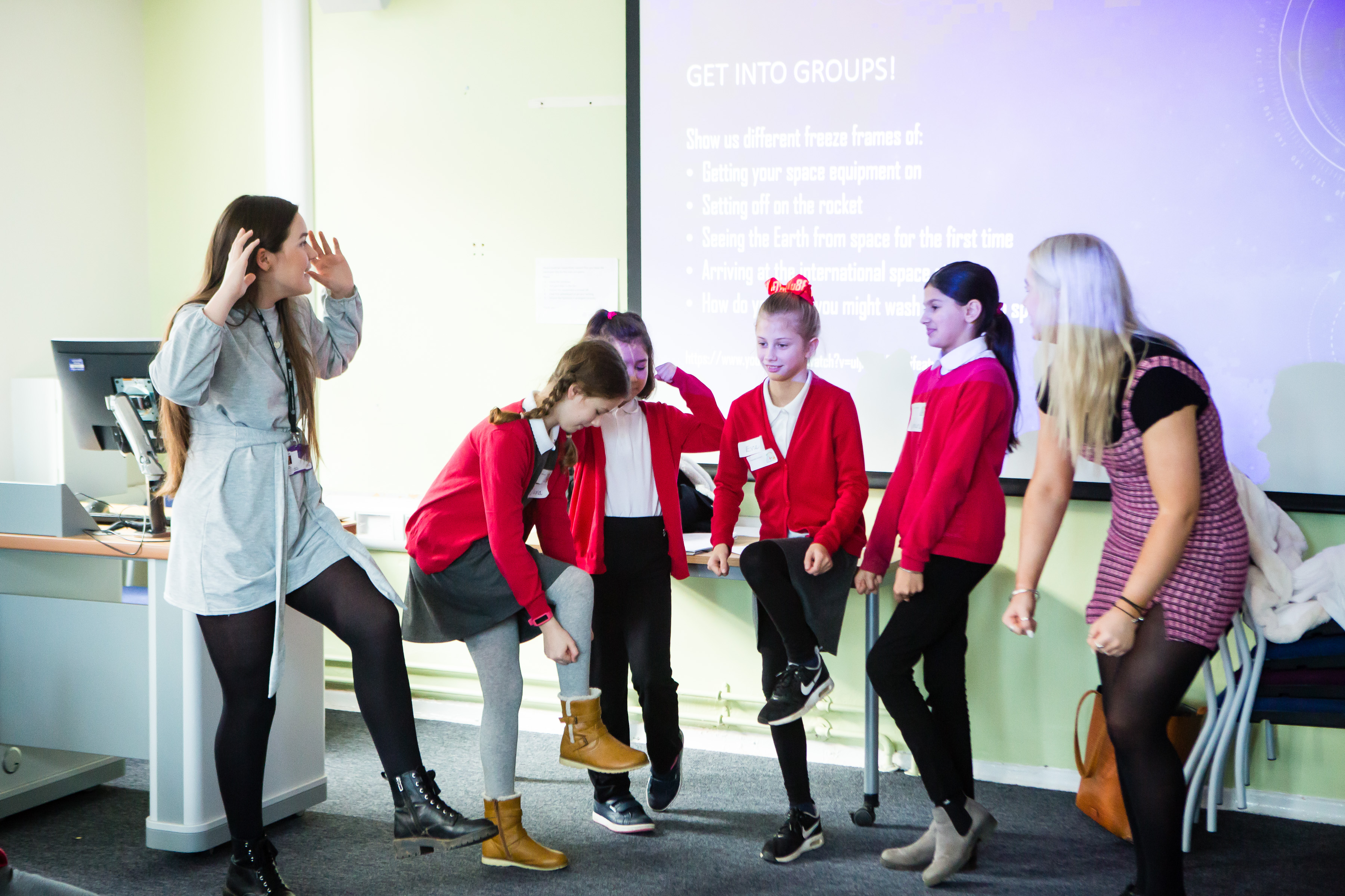 Children standing in front of a projector, making movements with their body, taking part in a creative activity. There is a teacher in the foreground. 