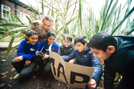 Pupils squatting on the ground, amongst long grass, looking at a piece of card that says 'map', some looking shocked and surprised, others are smiling. One girl has a camera around her neck. 