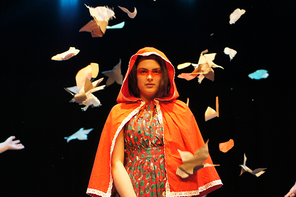 A young performer stands on a dark lit stage in a orange hooded cloak surrounded by paper origami birds floating around her.