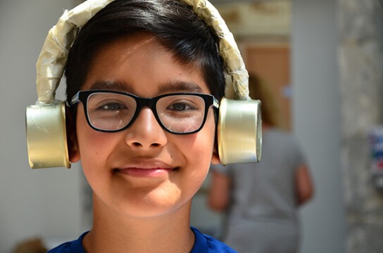 A young boy with black rimmed glasses smiles at the camera with a part of crafted gold foil headphones over his ears.